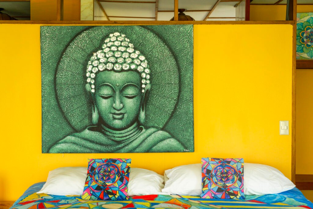 Green Buddha artwork above colorful bed in yellow room.